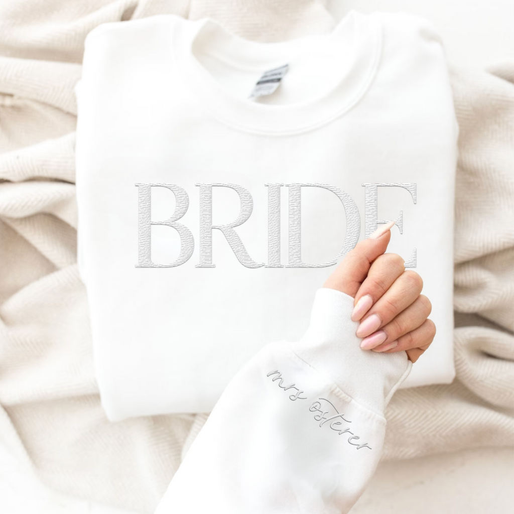 A white crewneck with 'BRIDE' embroidered on the front a a hand with a sleeve that says "mrs. osterer"