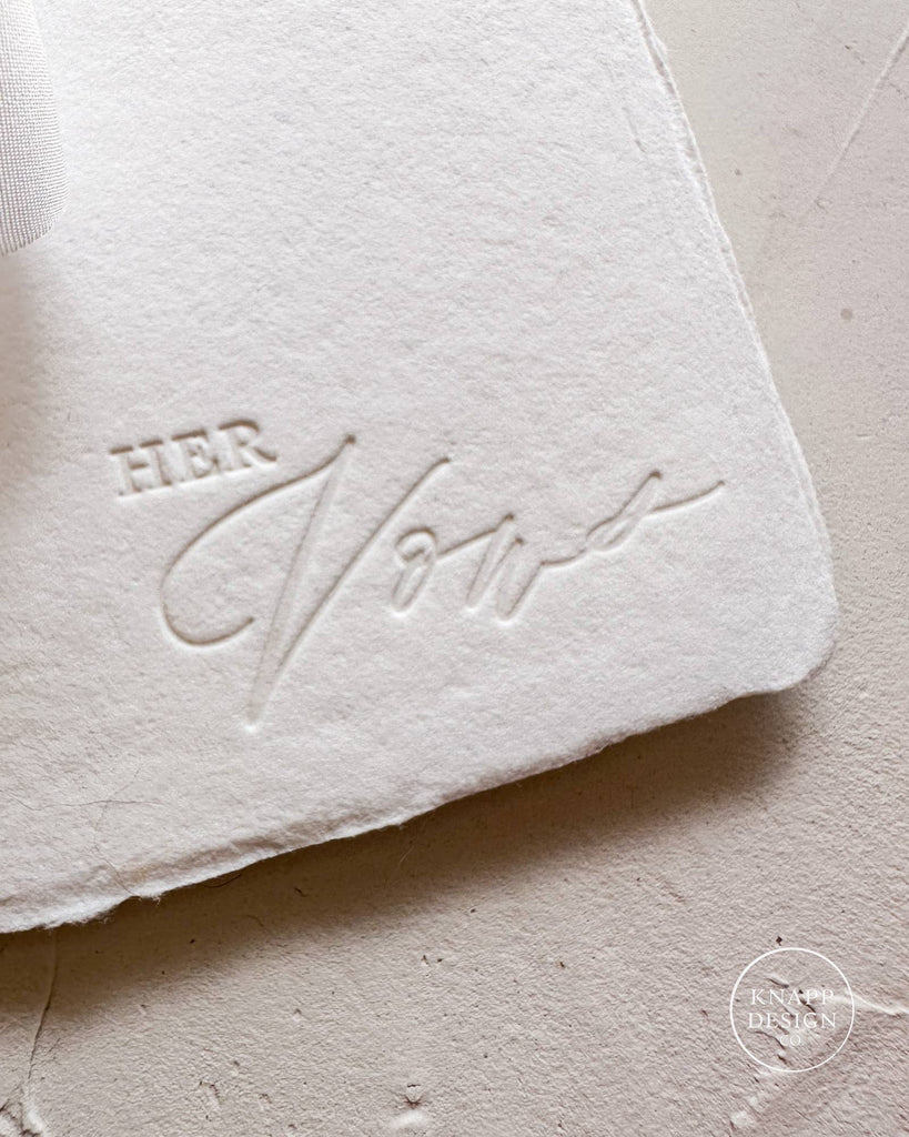 White vow book with white ribbon and "her vows" stamped on bottom right corner