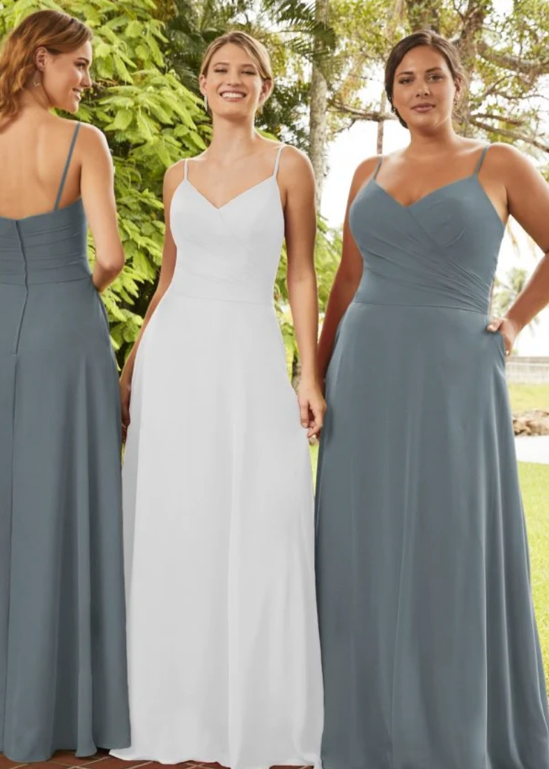 Three women wearing floor-length bridesmaid dresses outdoors. The center woman wears a white sleeveless V-neck gown, while the women on both sides wear matching dusty blue-gray spaghetti-strap floor-length gowns.