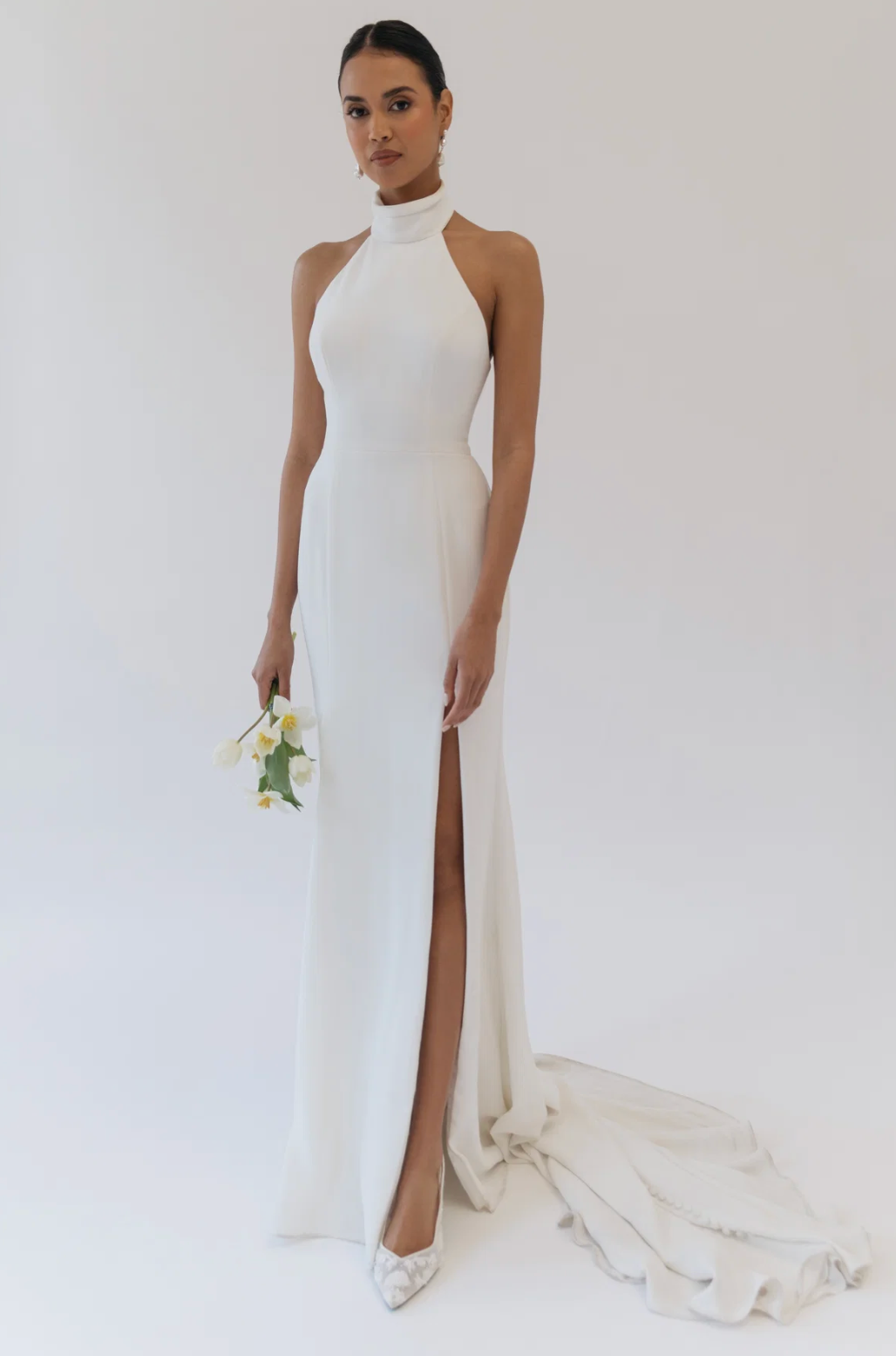 A woman stands in a studio wearing a floor-length white halter gown with a high thigh slit and a long train. She is holding a small bouquet of white flowers and wearing white pointed shoes against a plain light background.