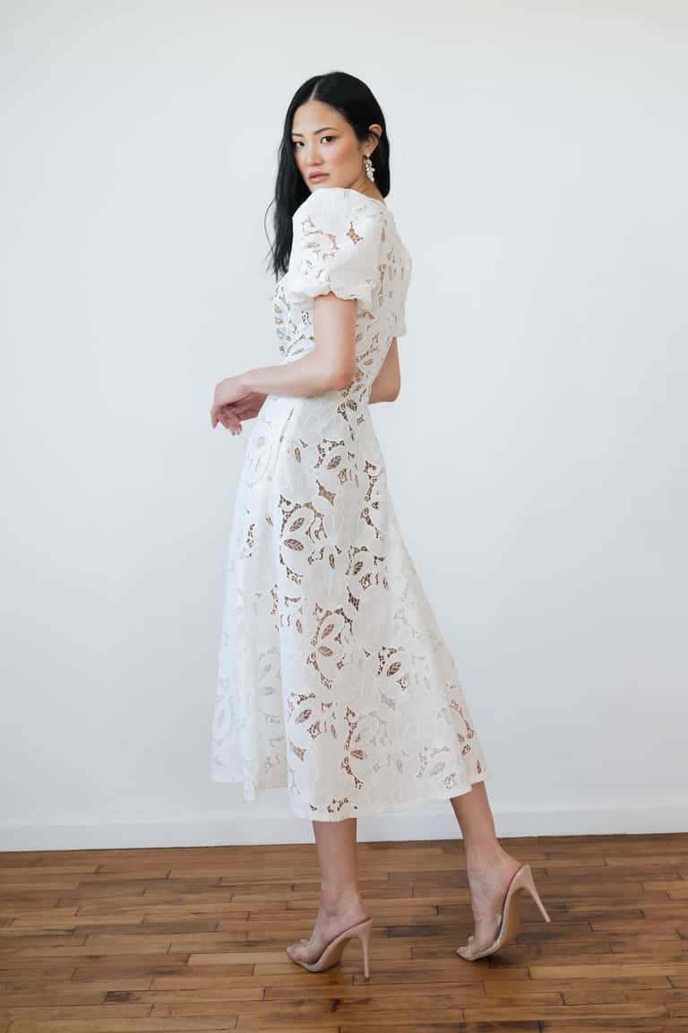 Woman wearing a white lace dress standing against a plain white wall.