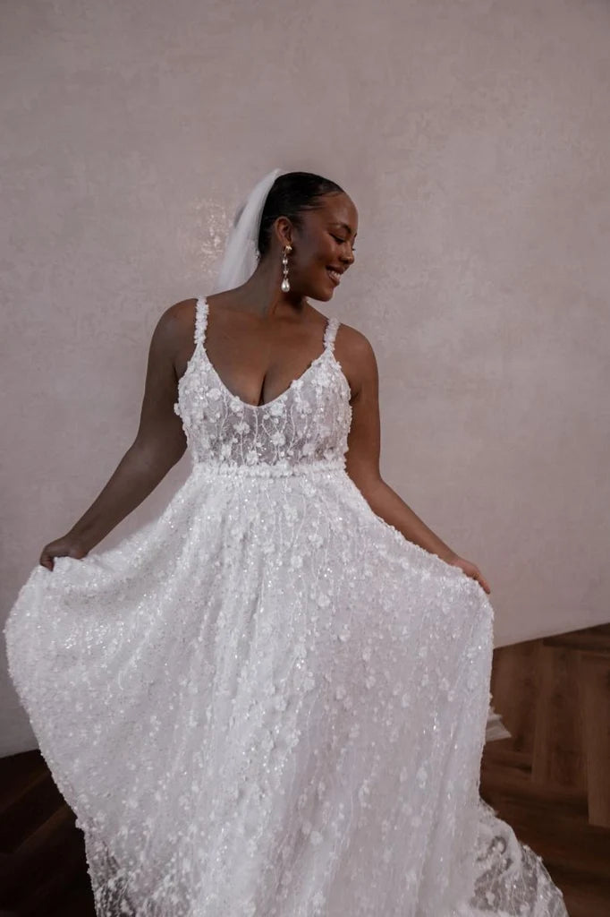 A woman wearing a white lace wedding dress with thin straps, a deep V-neckline, and a fitted bodice holding out the full skirt, photographed indoors against a textured beige wall. The dress is Lenni flowy by made with love