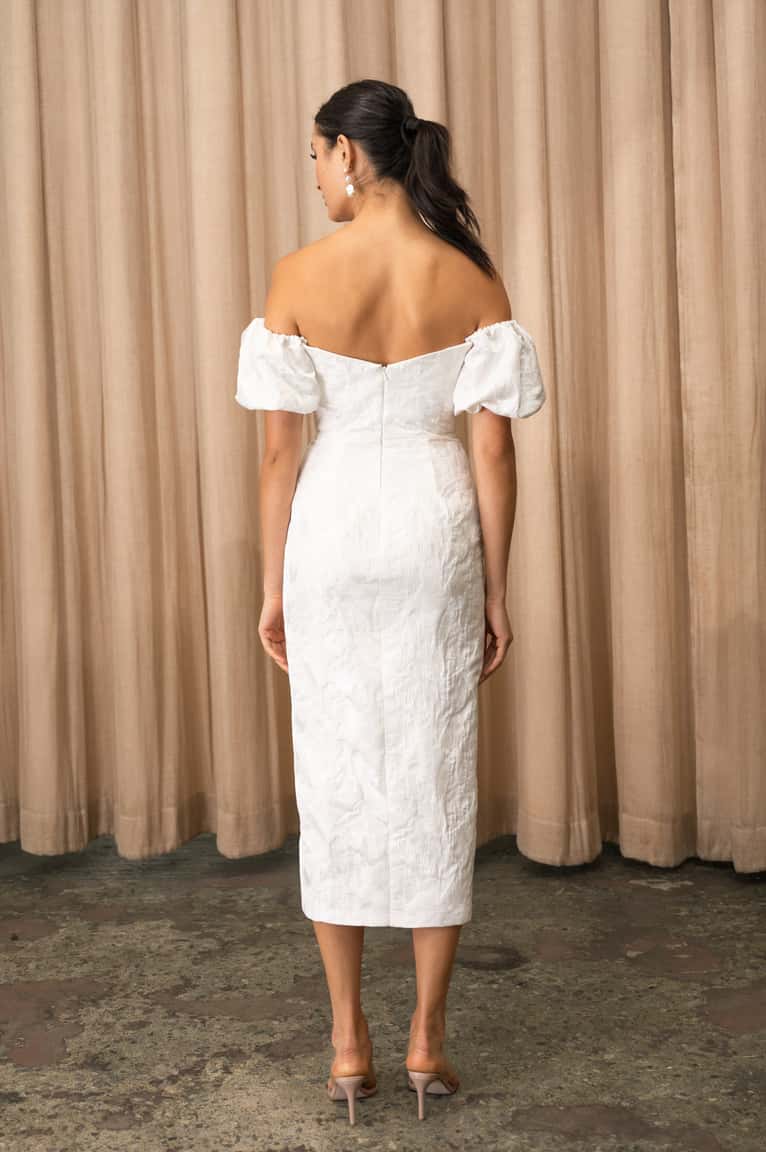 Woman wearing a white off-shoulder dress against a beige curtain background