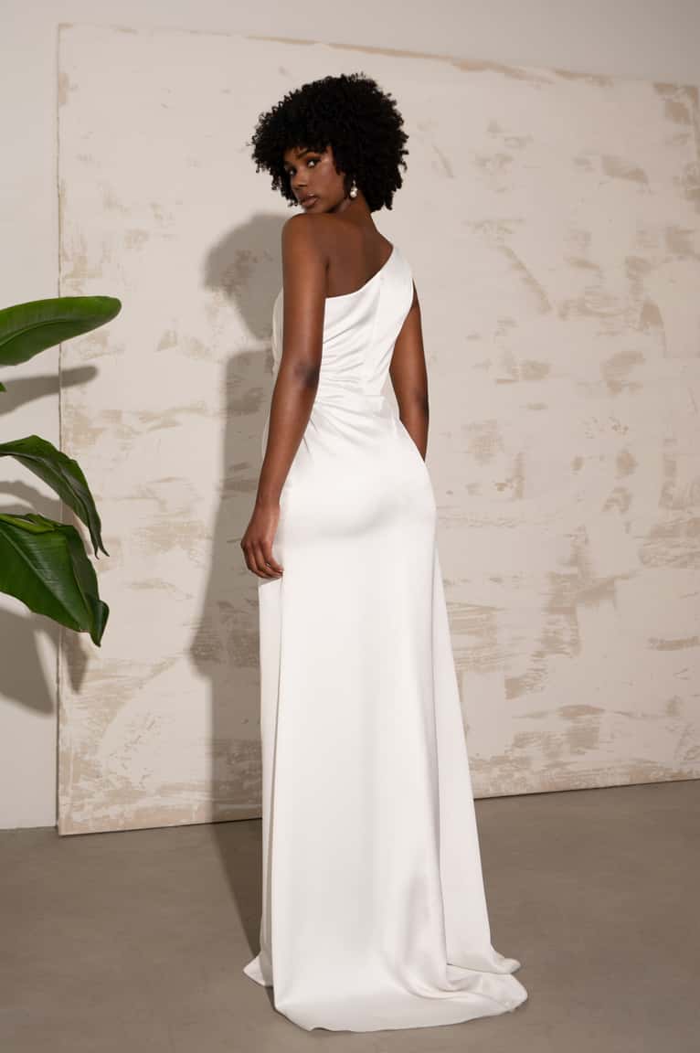 Woman in a white one-shoulder dress standing against a light-colored wall.