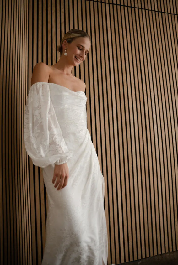 Woman in a white off-shoulder dress standing against a wooden paneled wall.