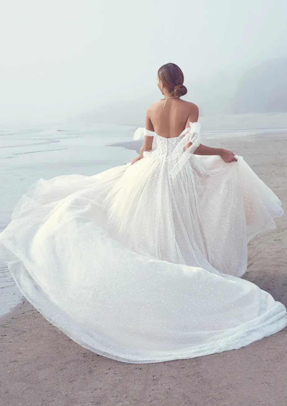 A bride in a voluminous white wedding gown sits on a beach, viewed from behind as she holds out the train of the dress. The gown has off-the-shoulder straps, a fitted lace-up back, and a long flowing train on a soft sandy shore with a misty ocean background.
