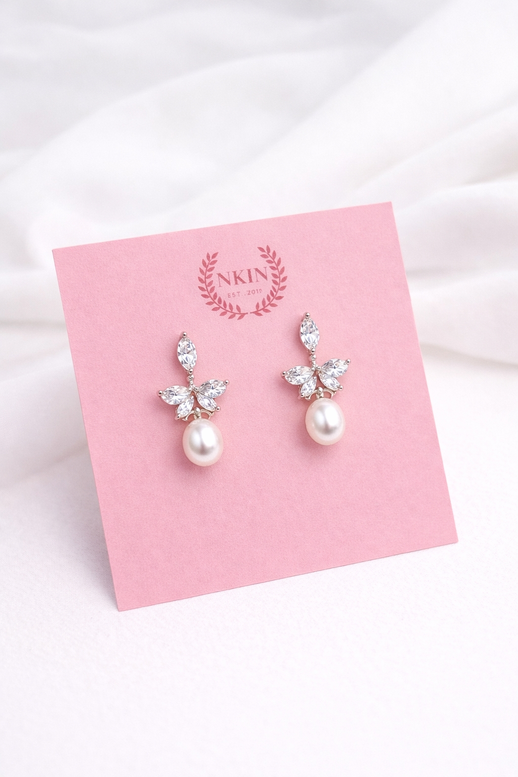 Pearl earrings with minimalist butterfly designs on a pink card with 'NKIN' branding.