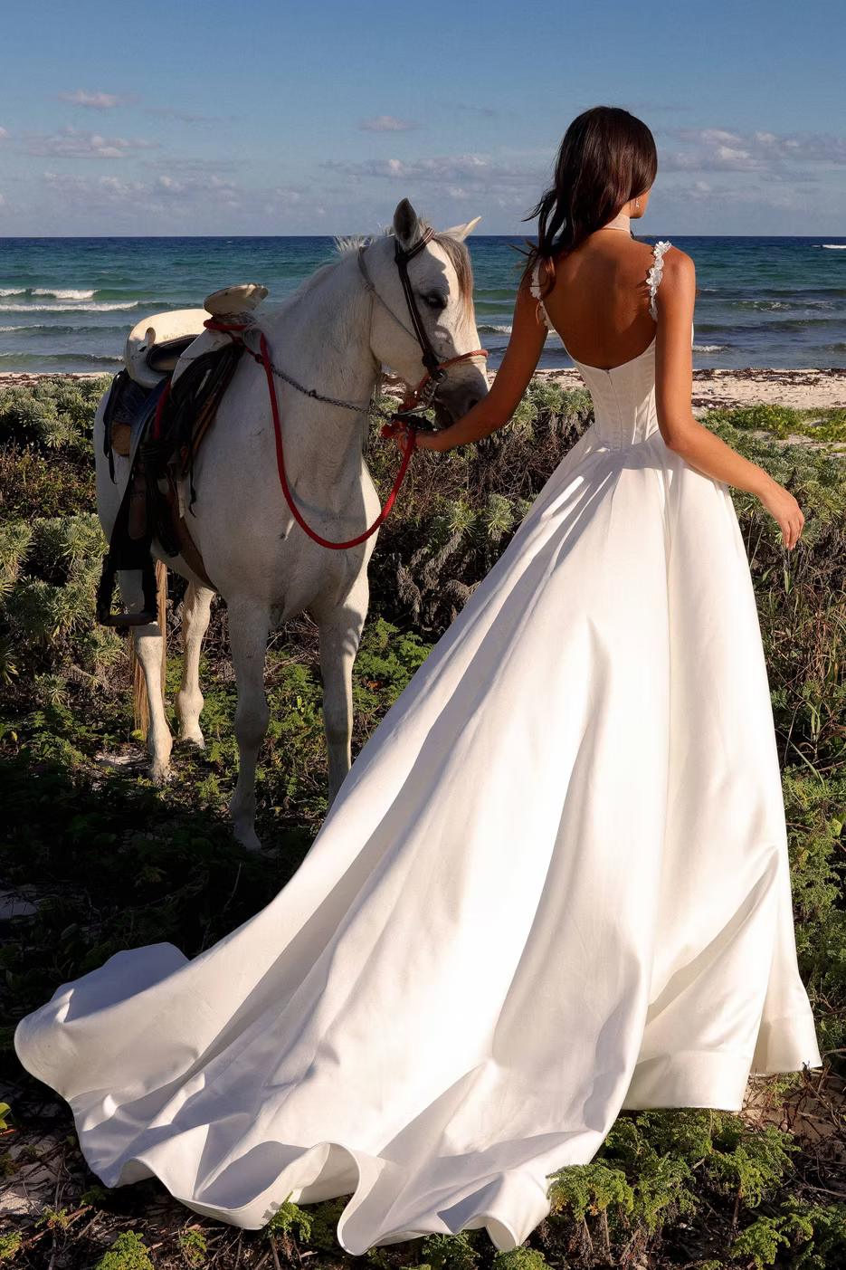 A woman in a flowing white wedding dress stands on a beach with a white horse beside her, photographed from behind to show the back of the dress and the ocean in the background.