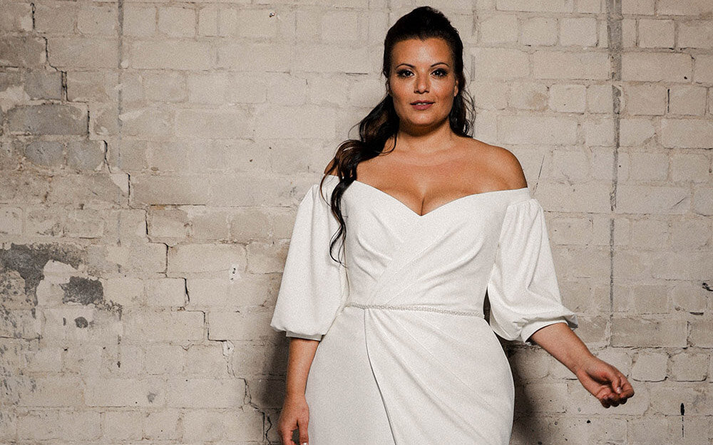 Closeup of a plus size model wearing Kim an off the shoulder puffy sleeved gown made of draped crepe.