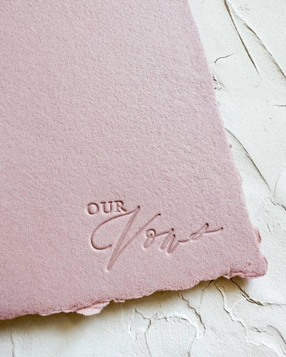 Pink vow book with white ribbon and "our vows" pressed in the bottom right corner