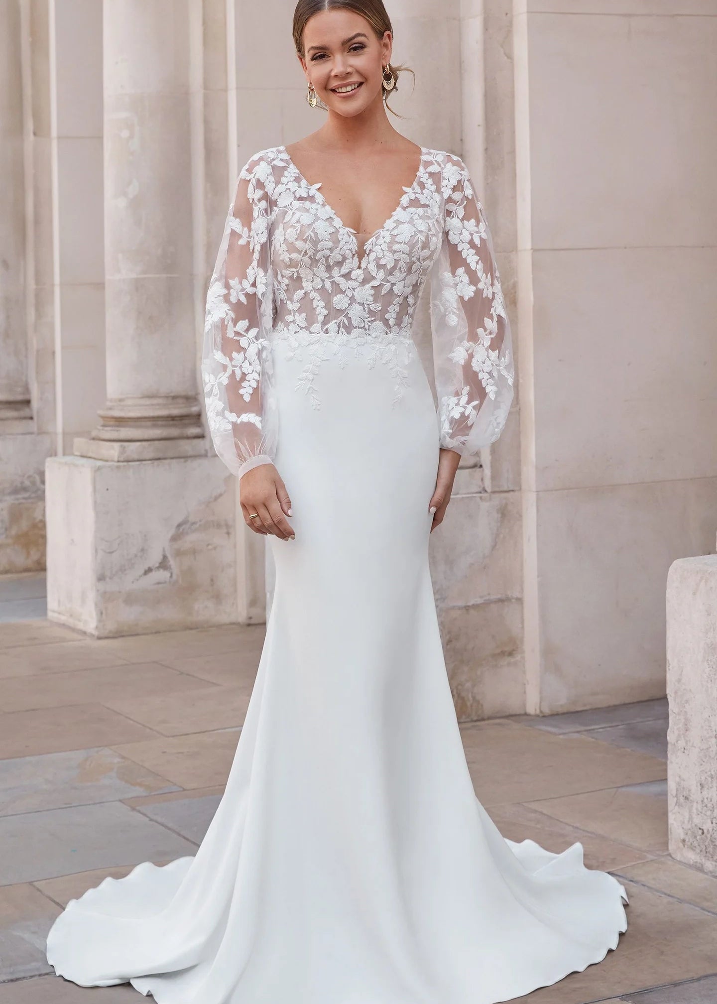 Woman wearing a white wedding dress with floral lace sleeves in an outdoor setting.