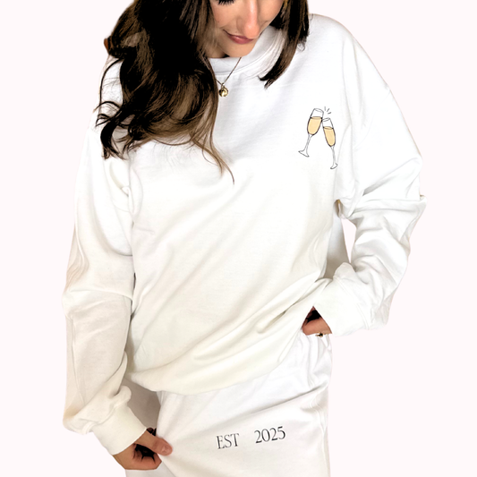 White sweatshirt with champagne glass design and 'EST 2025' text worn by a person on a white background