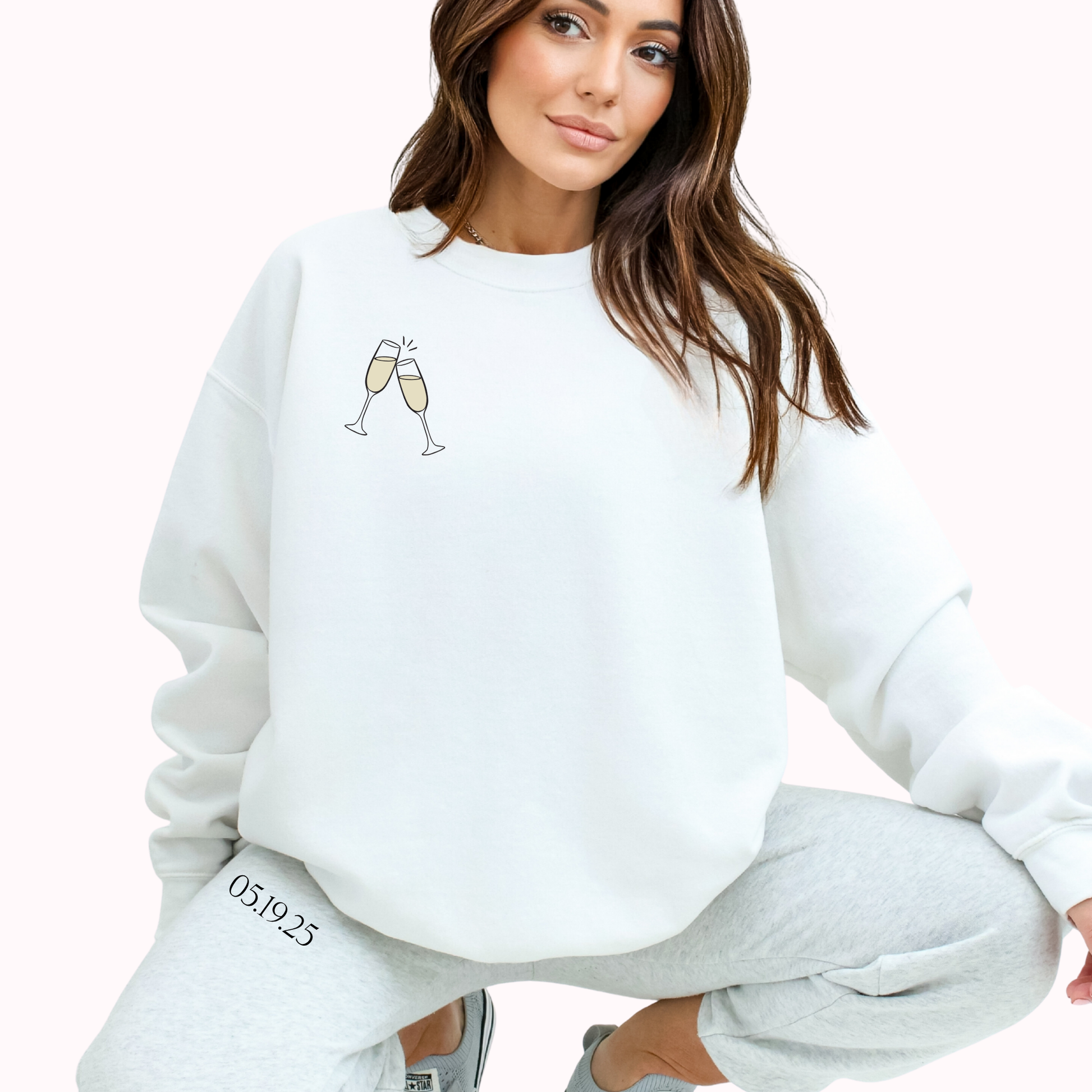 Person wearing a white sweatshirt with champagne glass design and text, sitting on a white background