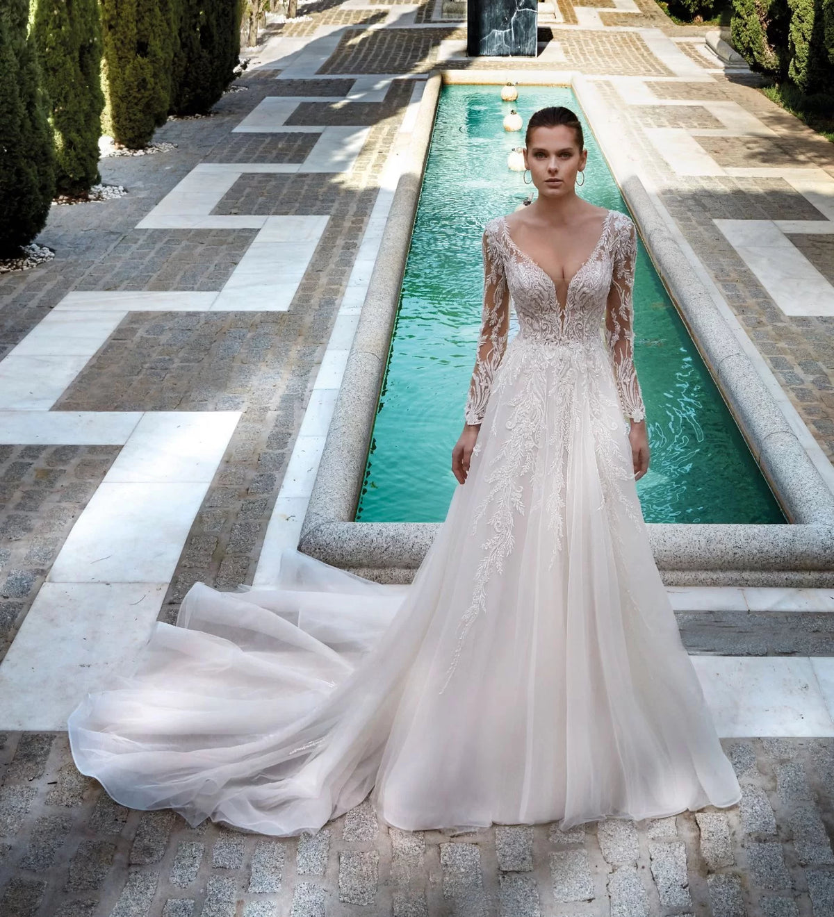 Model showing off 1193 in front of a long pool. A modern winter long sleeve wedding dress in tulle and lace with a fluid silhouette and an open back