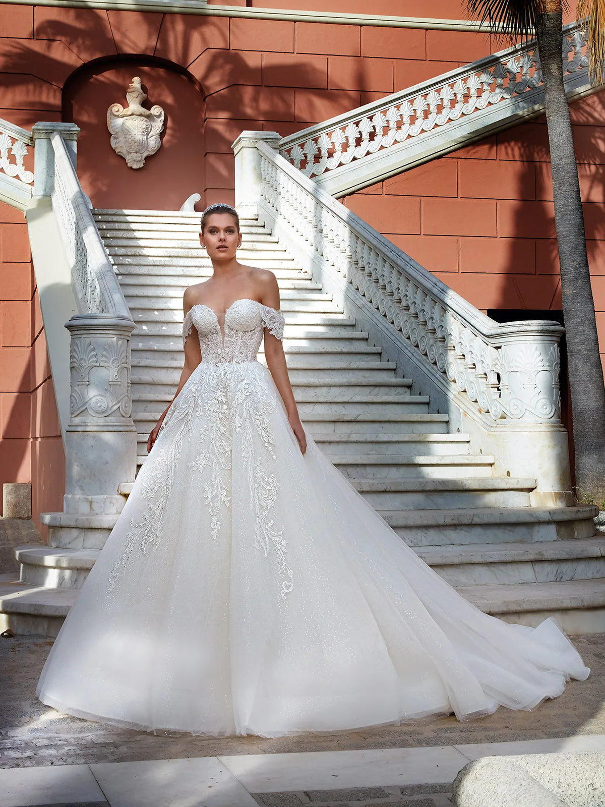 A woman wearing an off-the-shoulder white ball gown with floral lace embroidery stands on a grand outdoor staircase in front of a terracotta-colored building.