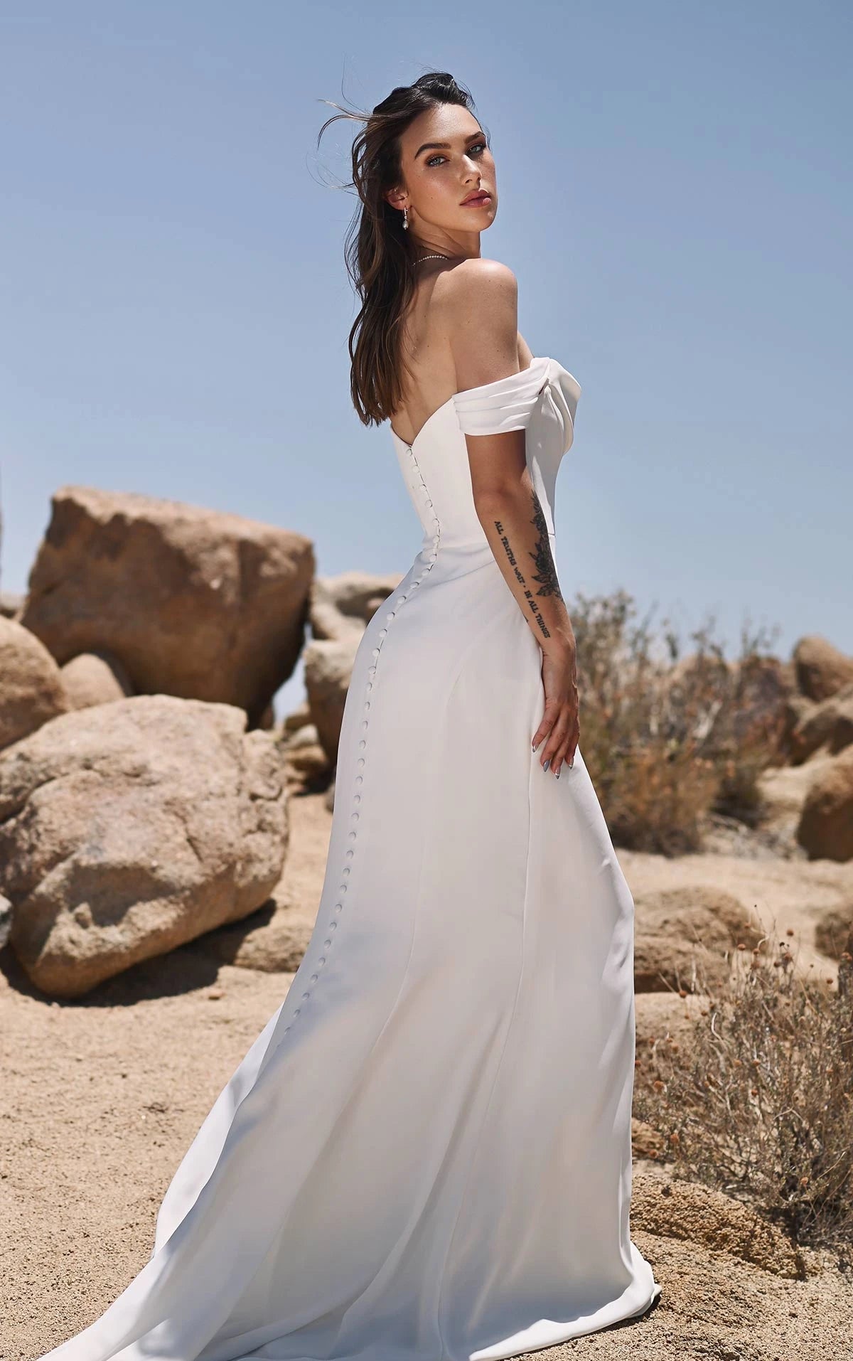 A woman wearing a white off-the-shoulder wedding gown stands outdoors in a rocky desert landscape under a clear blue sky, photographed from the back as the dress flows behind her.