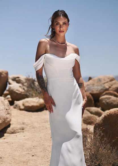 A woman wearing a white off-the-shoulder floor-length gown stands outdoors in a rocky desert landscape under a clear blue sky.
