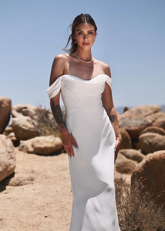 A woman wearing a white off-the-shoulder floor-length gown stands outdoors in a rocky desert landscape under a clear blue sky.