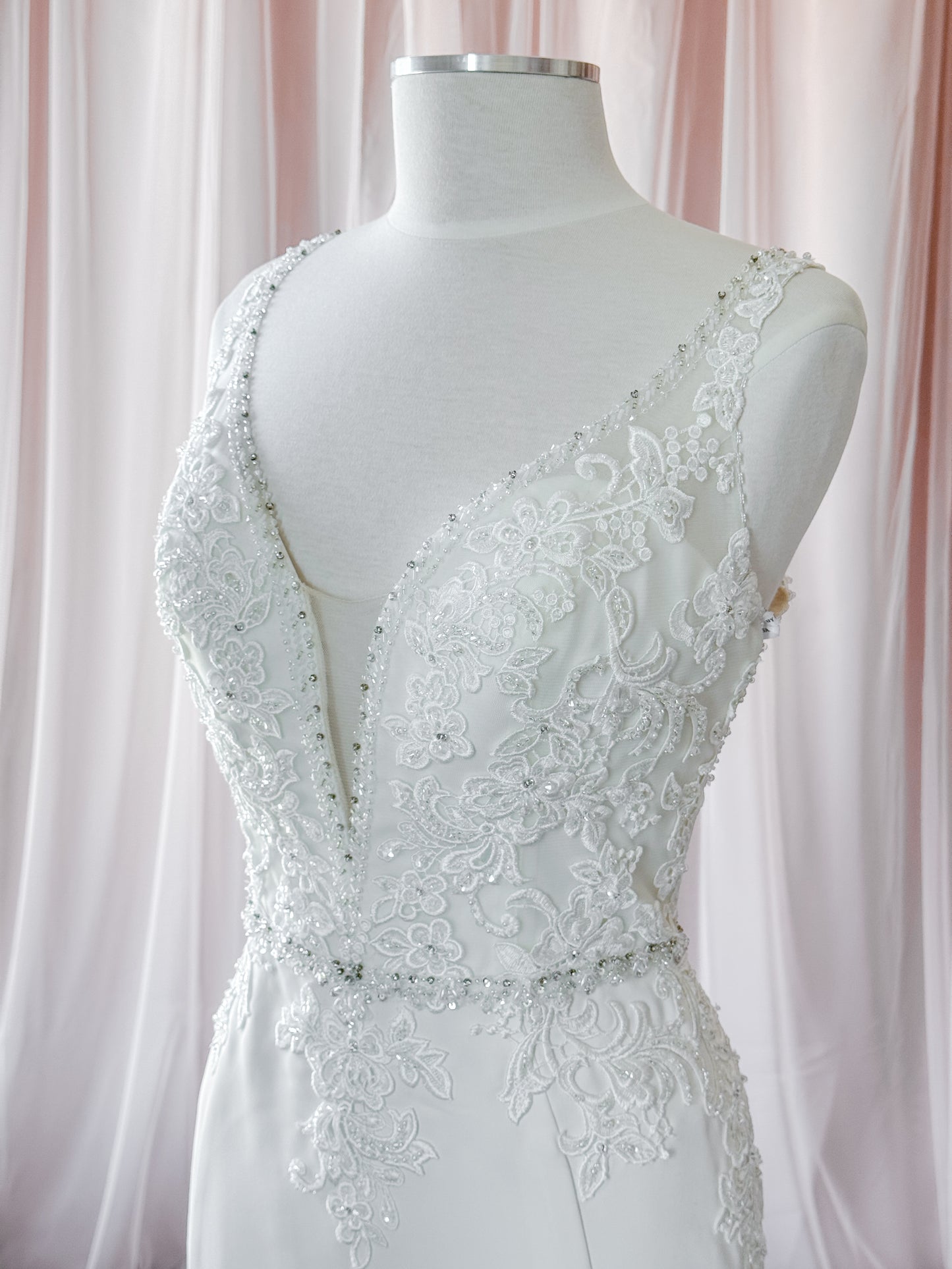 White lace wedding dress by Kenneth Winston displayed on a white mannequin torso, featuring a deep V-neckline, beaded embellishments, and floral lace detailing against a soft pink draped fabric background at Never knew I Needed Bridal Outlet