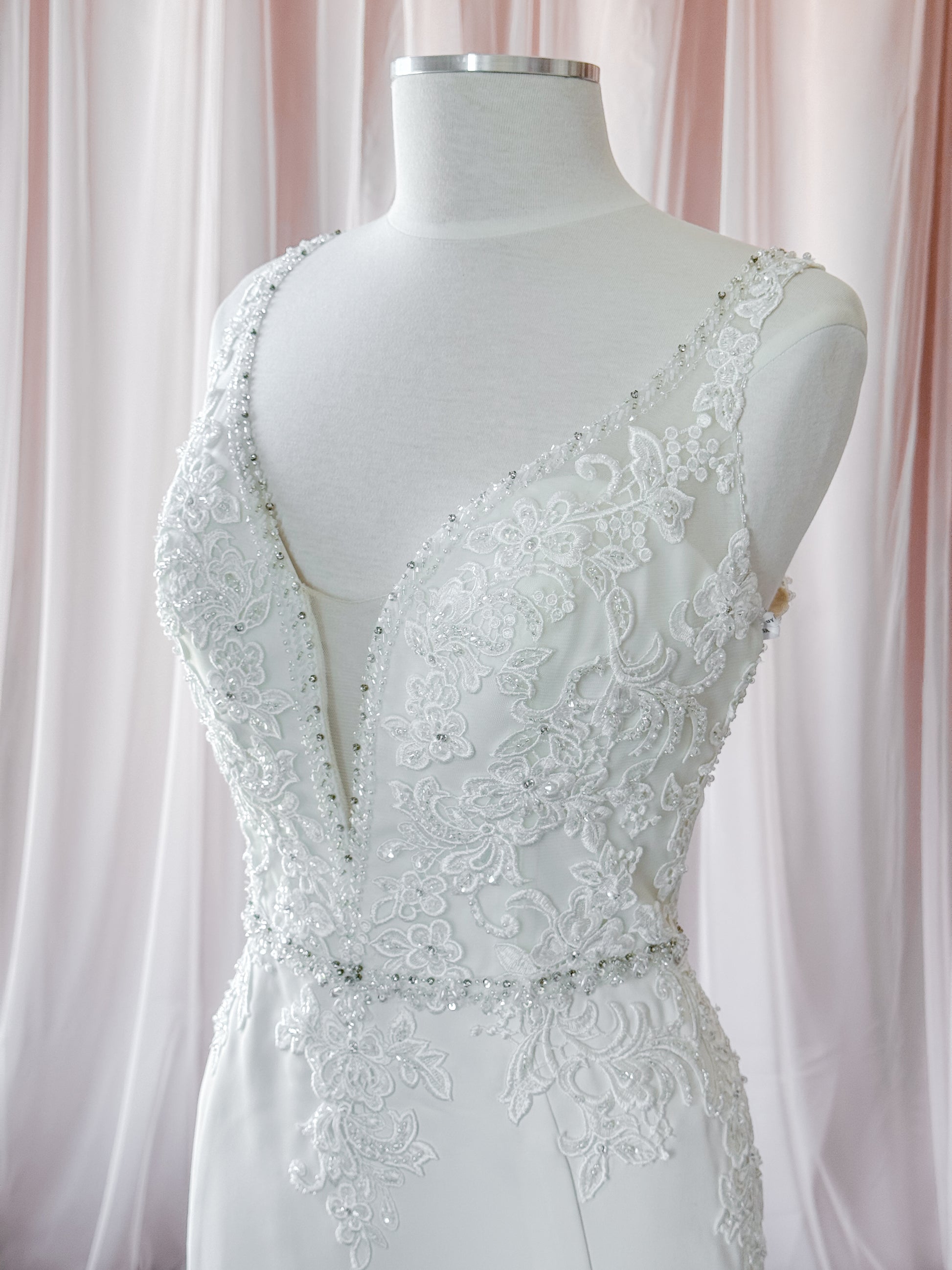 White lace wedding dress by Kenneth Winston displayed on a white mannequin torso, featuring a deep V-neckline, beaded embellishments, and floral lace detailing against a soft pink draped fabric background at Never knew I Needed Bridal Outlet