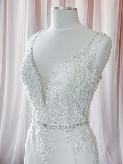 White lace wedding dress by Kenneth Winston displayed on a white mannequin torso, featuring a deep V-neckline, beaded embellishments, and floral lace detailing against a soft pink draped fabric background at Never knew I Needed Bridal Outlet