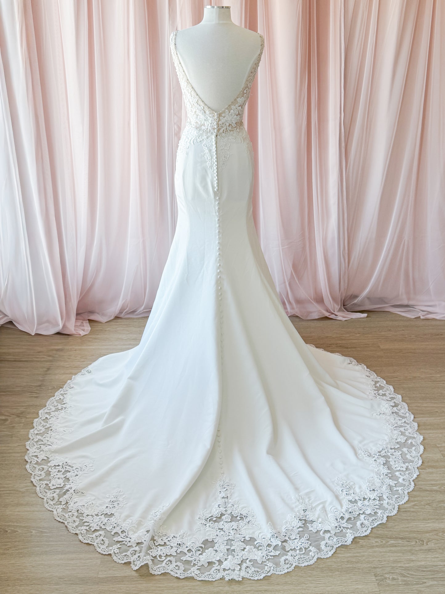 A white bridal gown displayed on a mannequin torso, featuring a deep V backline, lace bodice, and a long train with a scalloped lace hem, set against a the NKIN Ottawa Bridal backdrop of soft pink draped fabric and light wood flooring
