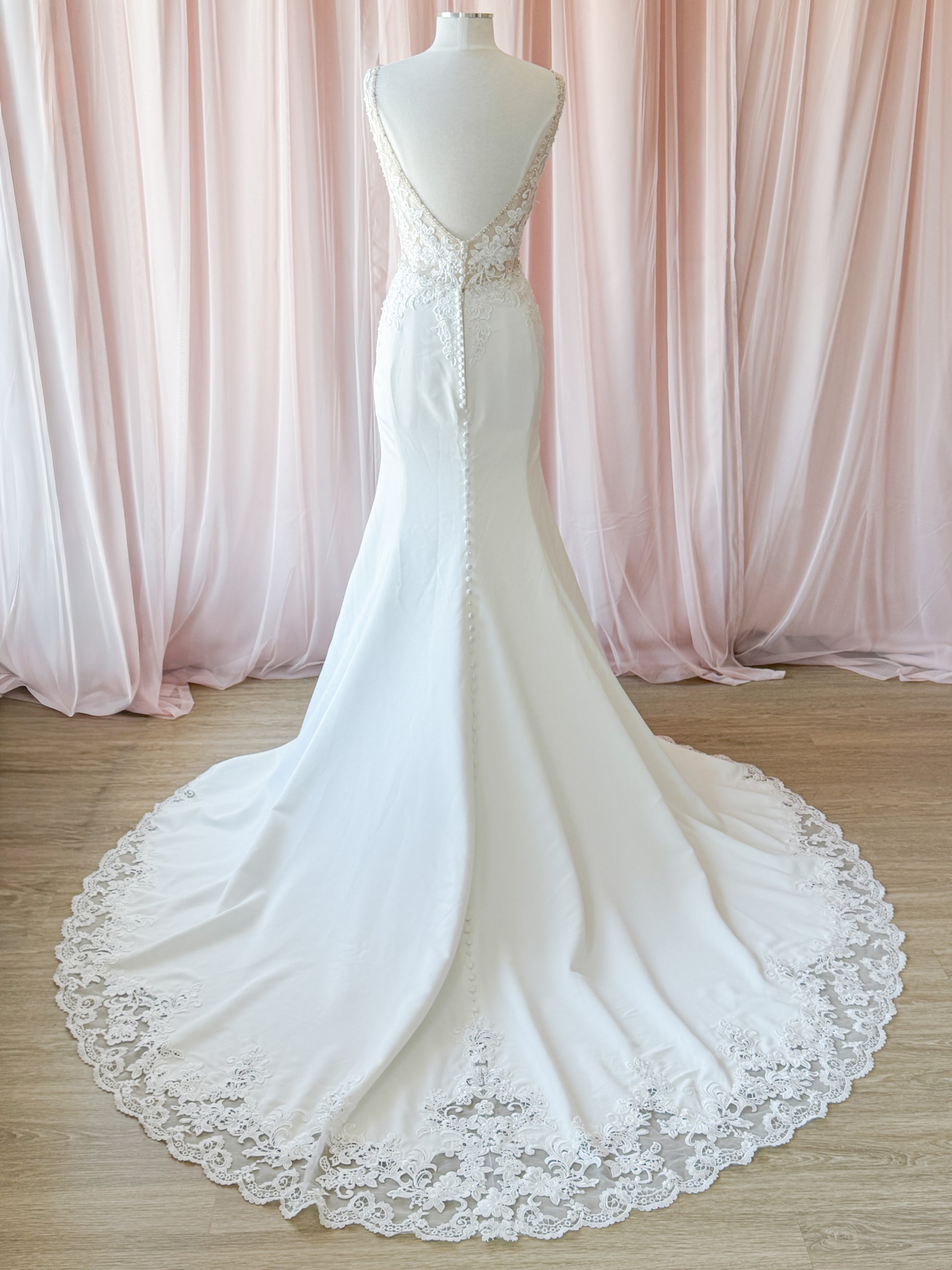 A white bridal gown displayed on a mannequin torso, featuring a deep V backline, lace bodice, and a long train with a scalloped lace hem, set against a the NKIN Ottawa Bridal backdrop of soft pink draped fabric and light wood flooring