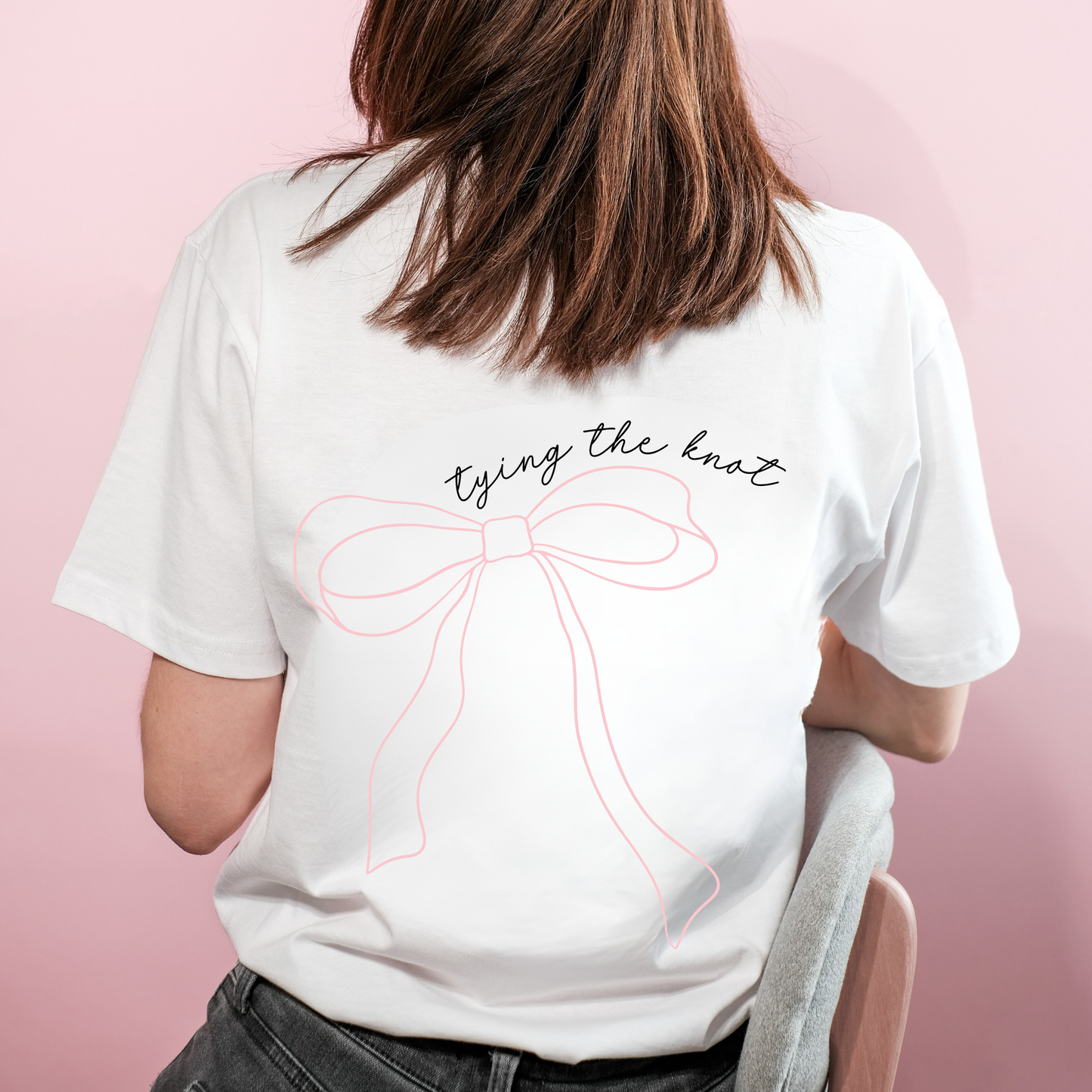 Person wearing a white t-shirt with a bow design and text on the back against a pink background
