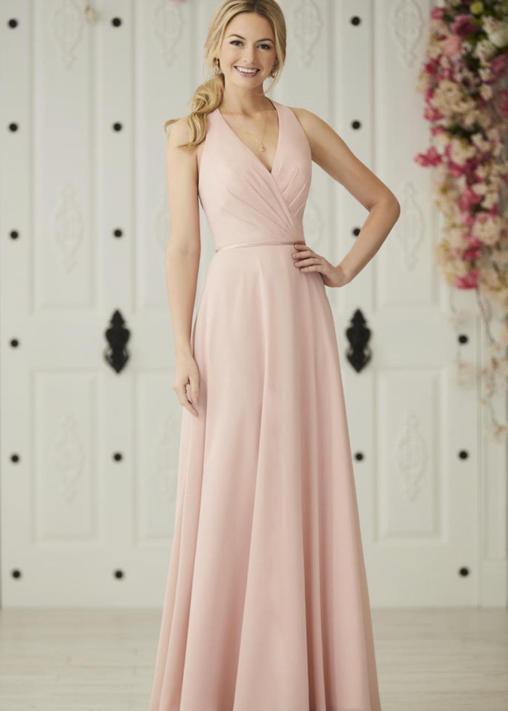 A woman modeling a floor-length blush pink formal gown with a V-neckline and sleeveless design, standing in front of a white decorative door with floral arrangements.