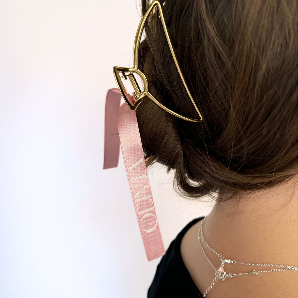 A gold claw clip with a pink ribbon customized with "olivia" in white a brunette girl's hair