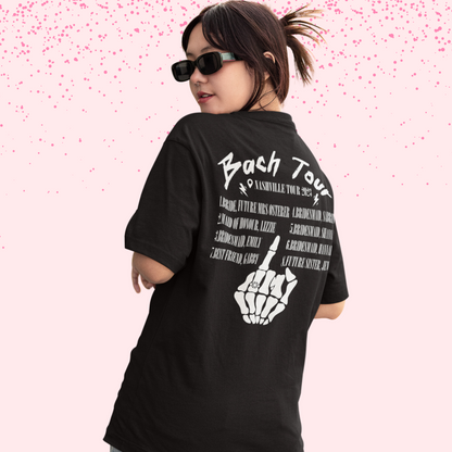 Young woman wearing a black shirt that says "bach tour" in white with names and a skeleton hand holding up their fourth finger