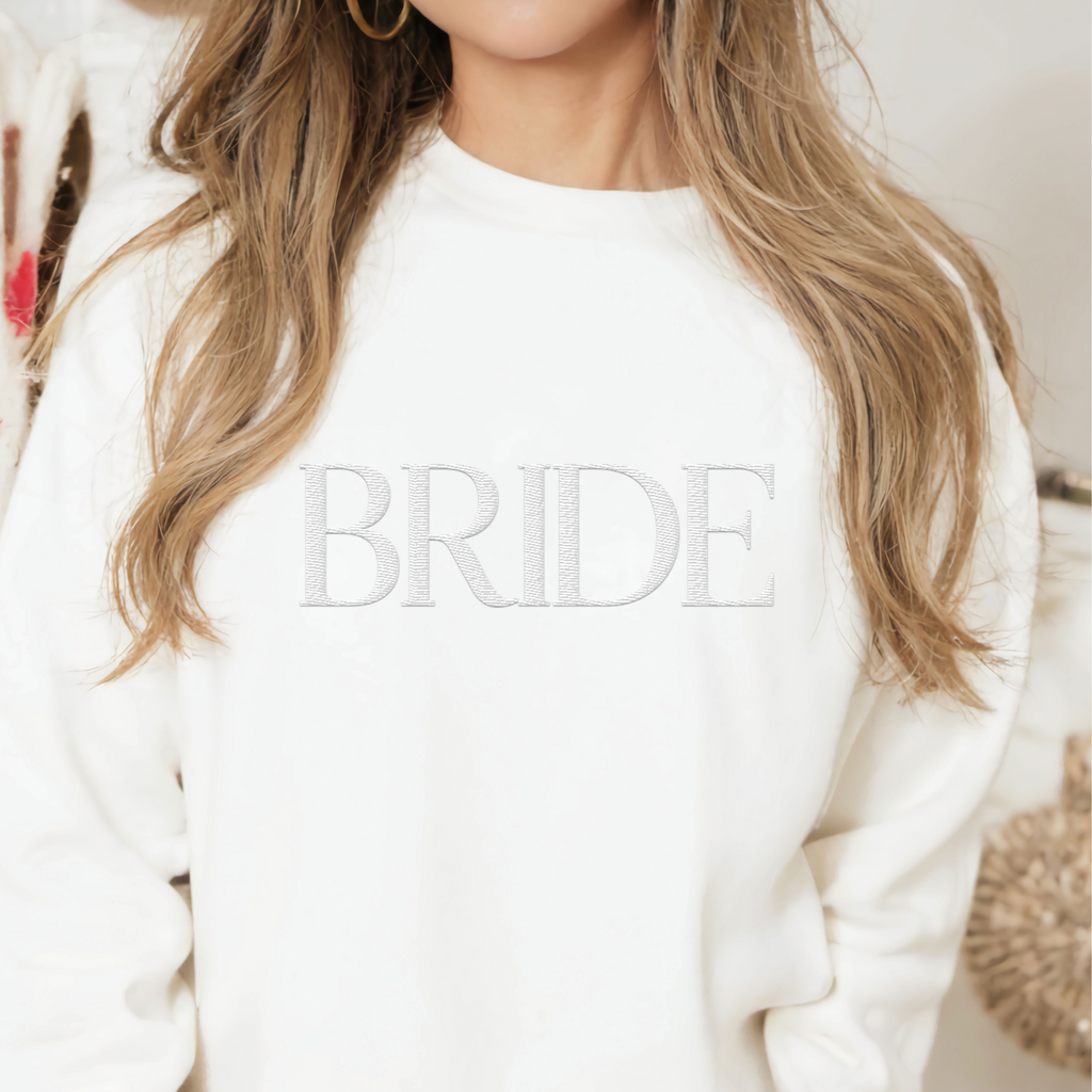 A young woman wearing a white crewneck with "BRIDE" embroidered on the front.