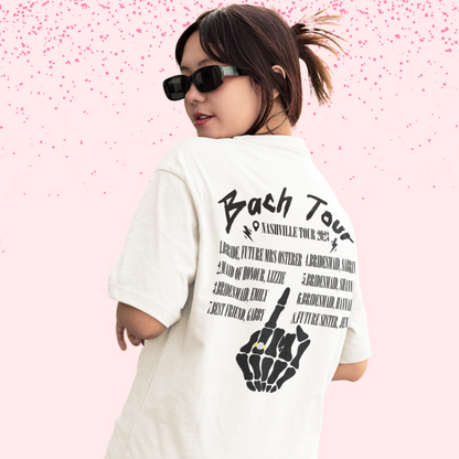 Young woman wearing a white shirt that says "bach tour" in white with names and a skeleton hand holding up their fourth finger