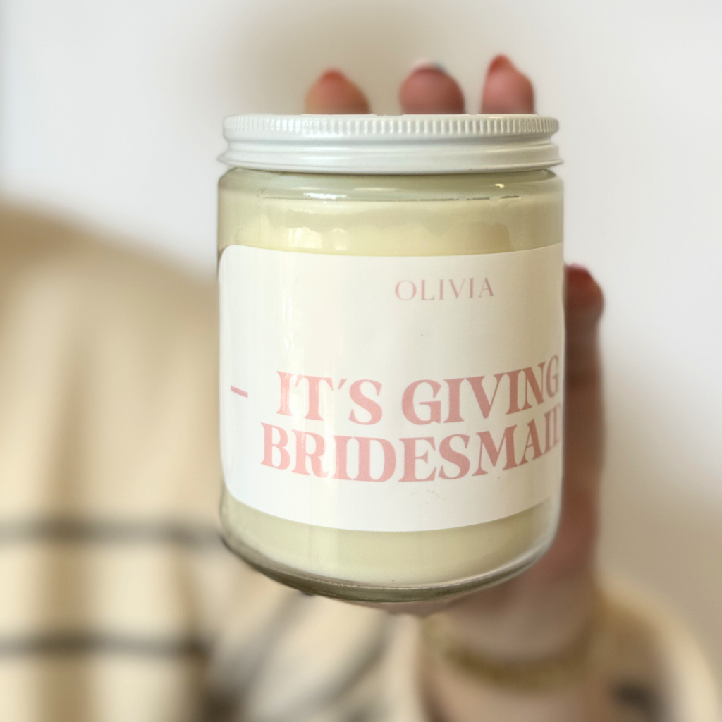 A white jarred candle with "Olivia - Its Giving Bridesmaid" written in pink