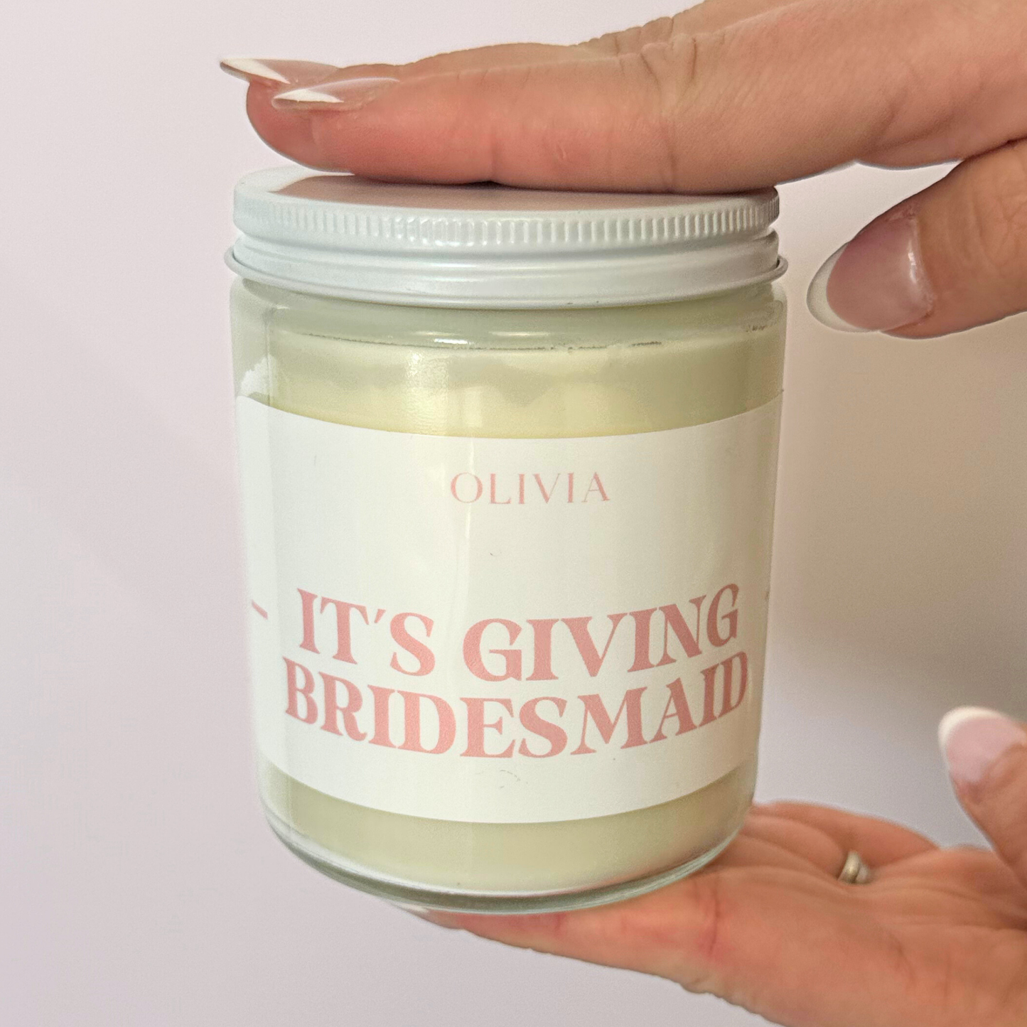 A white jarred candle with "Olivia - Its Giving Bridesmaid" written in pink