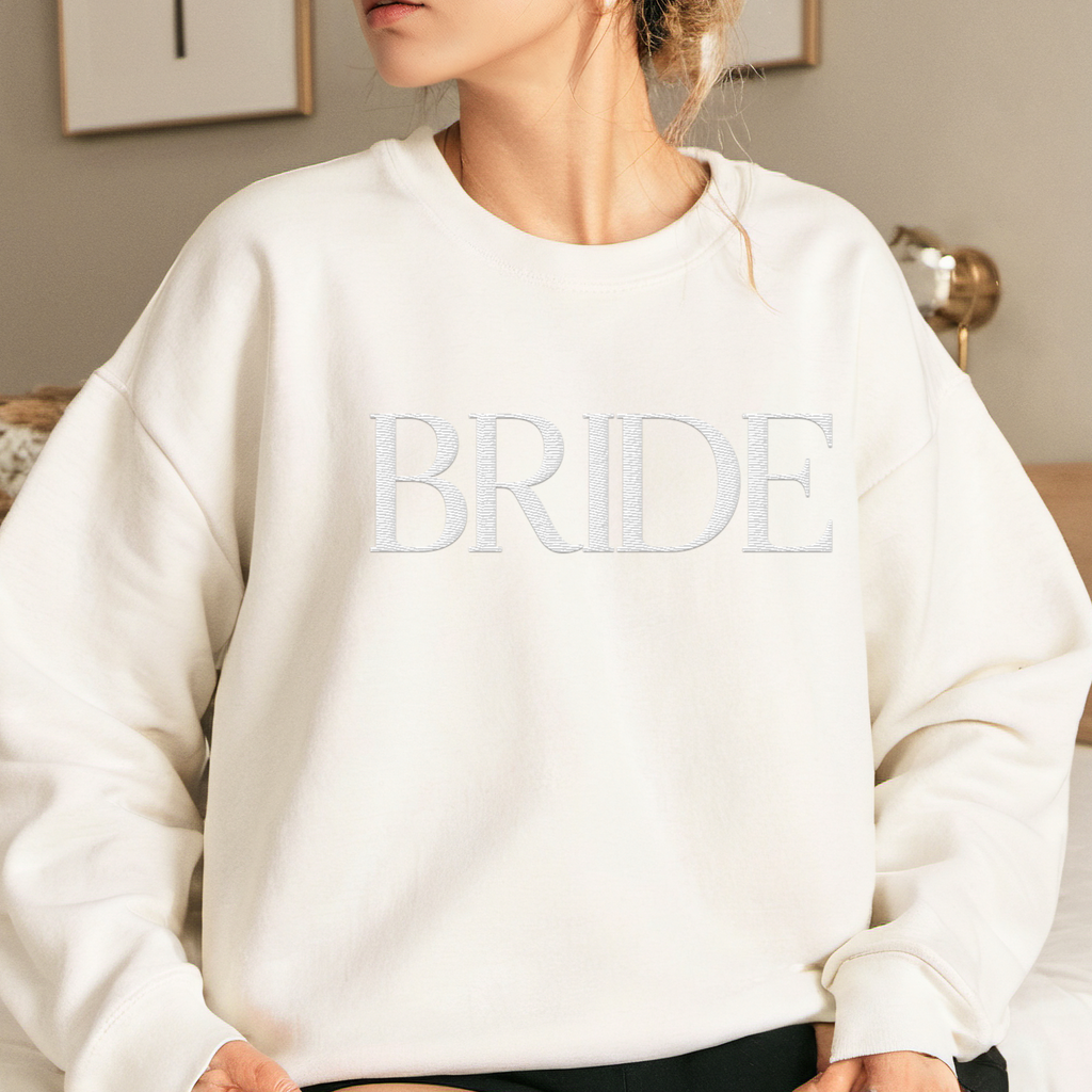 A young woman wearing a white crewneck with "BRIDE" embroidered on the front.