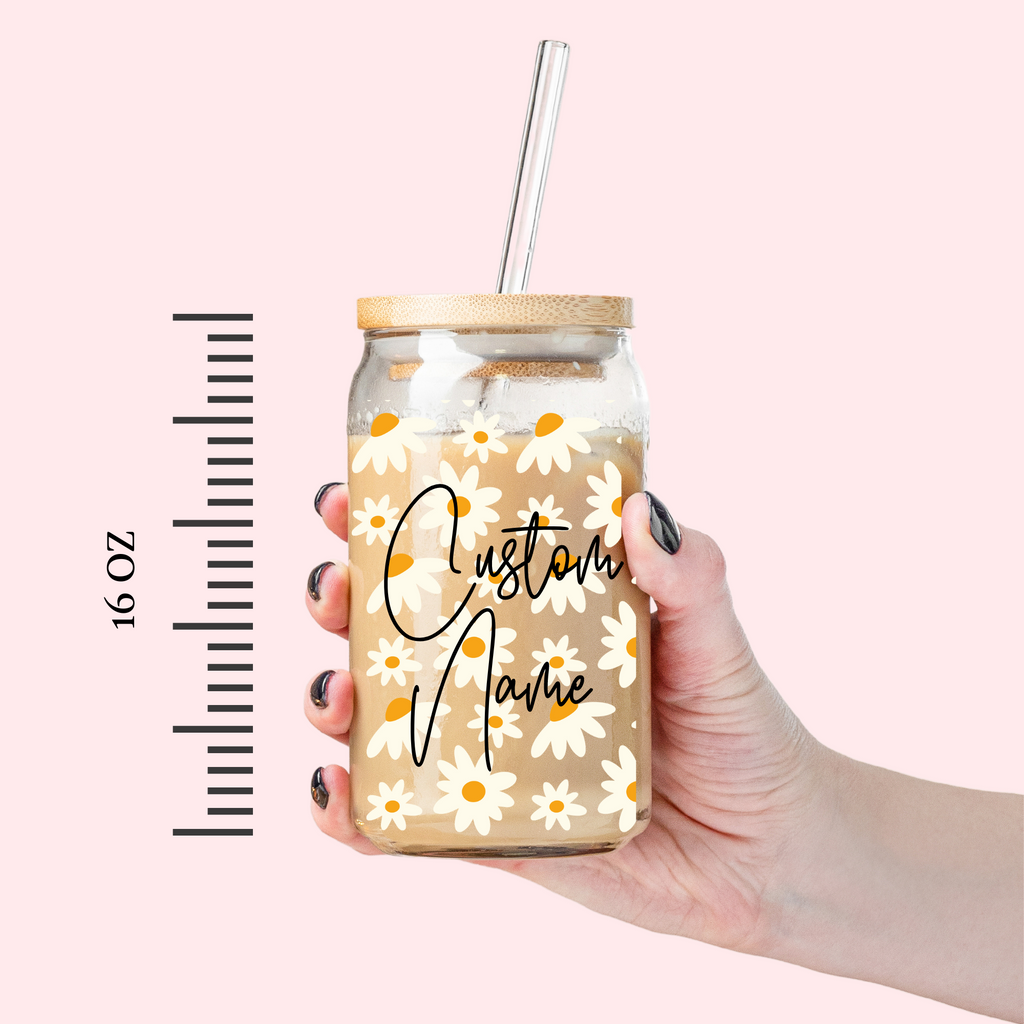 Glass tumbler with "custom name" written on it with daisy florals held by a hand. "16 oz" placed on the side