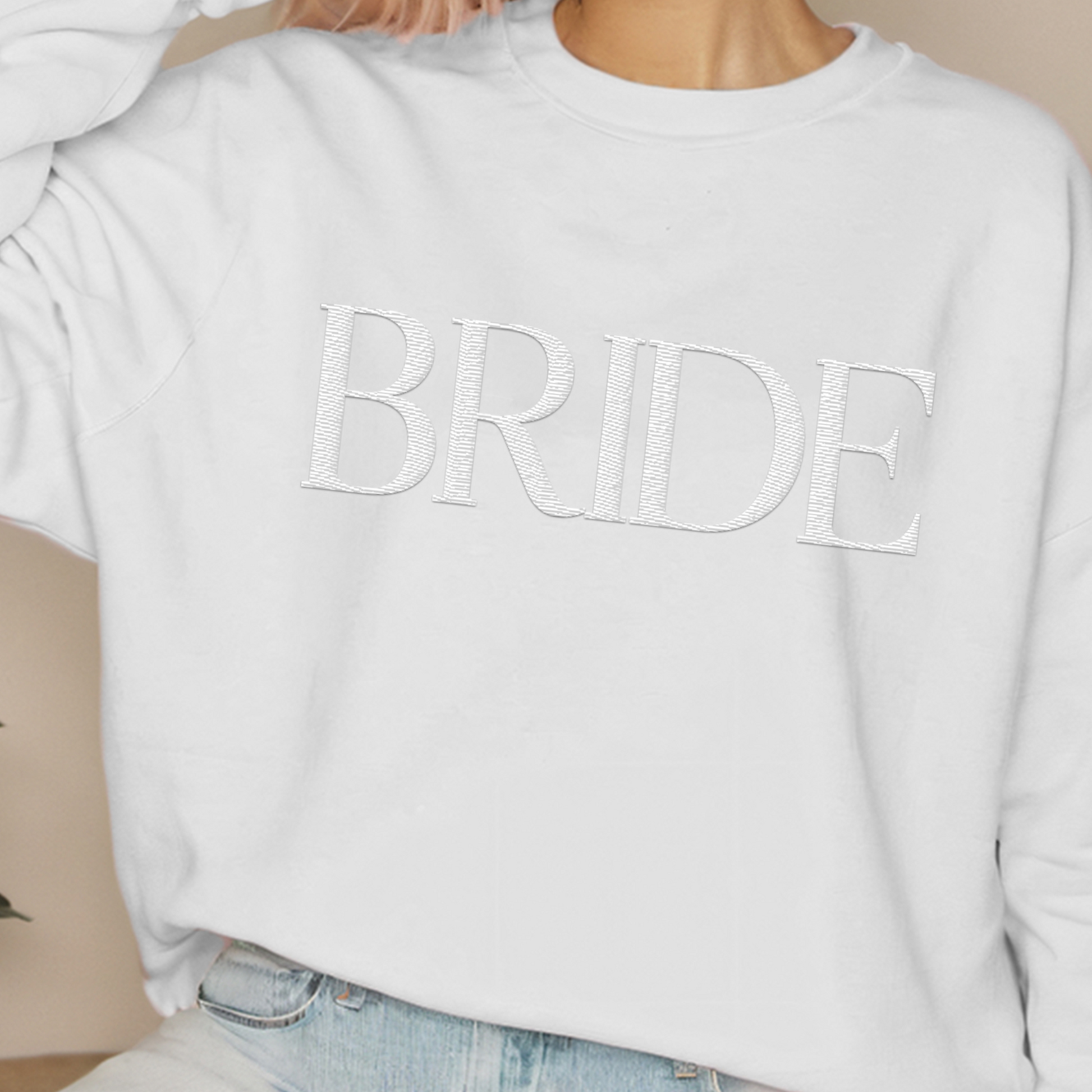 A young woman wearing a white crewneck with "BRIDE" embroidered on the front.