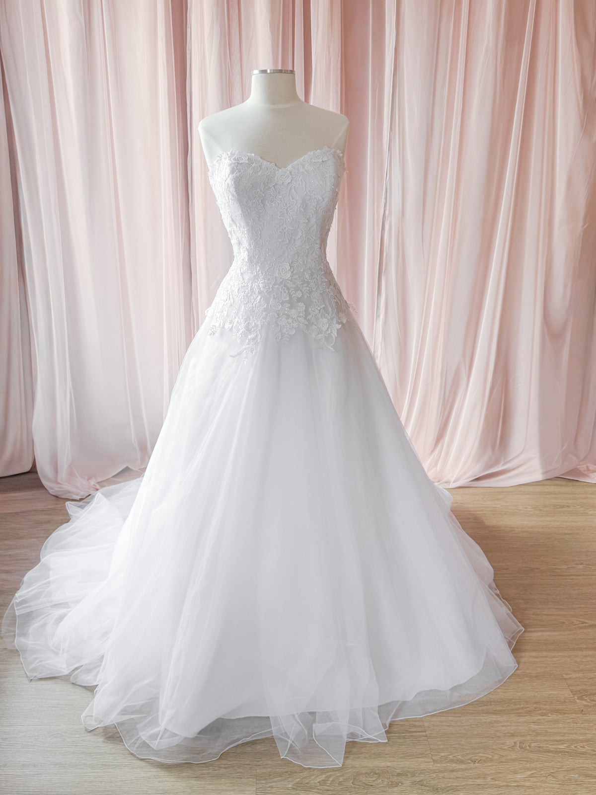 A white strapless wedding gown with a sweetheart neckline, lace-embellished bodice, and full tulle skirt displayed on a mannequin in front of soft pink draped fabric and a light wood floor.