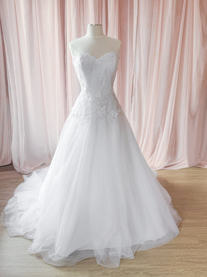 A white strapless wedding gown with a sweetheart neckline, lace-embellished bodice, and full tulle skirt displayed on a mannequin in front of soft pink draped fabric and a light wood floor.
