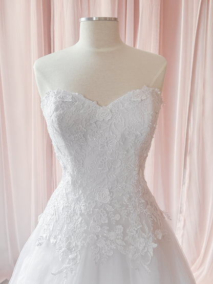 White strapless lace wedding dress displayed on a cream mannequin torso against soft pink draped fabric.