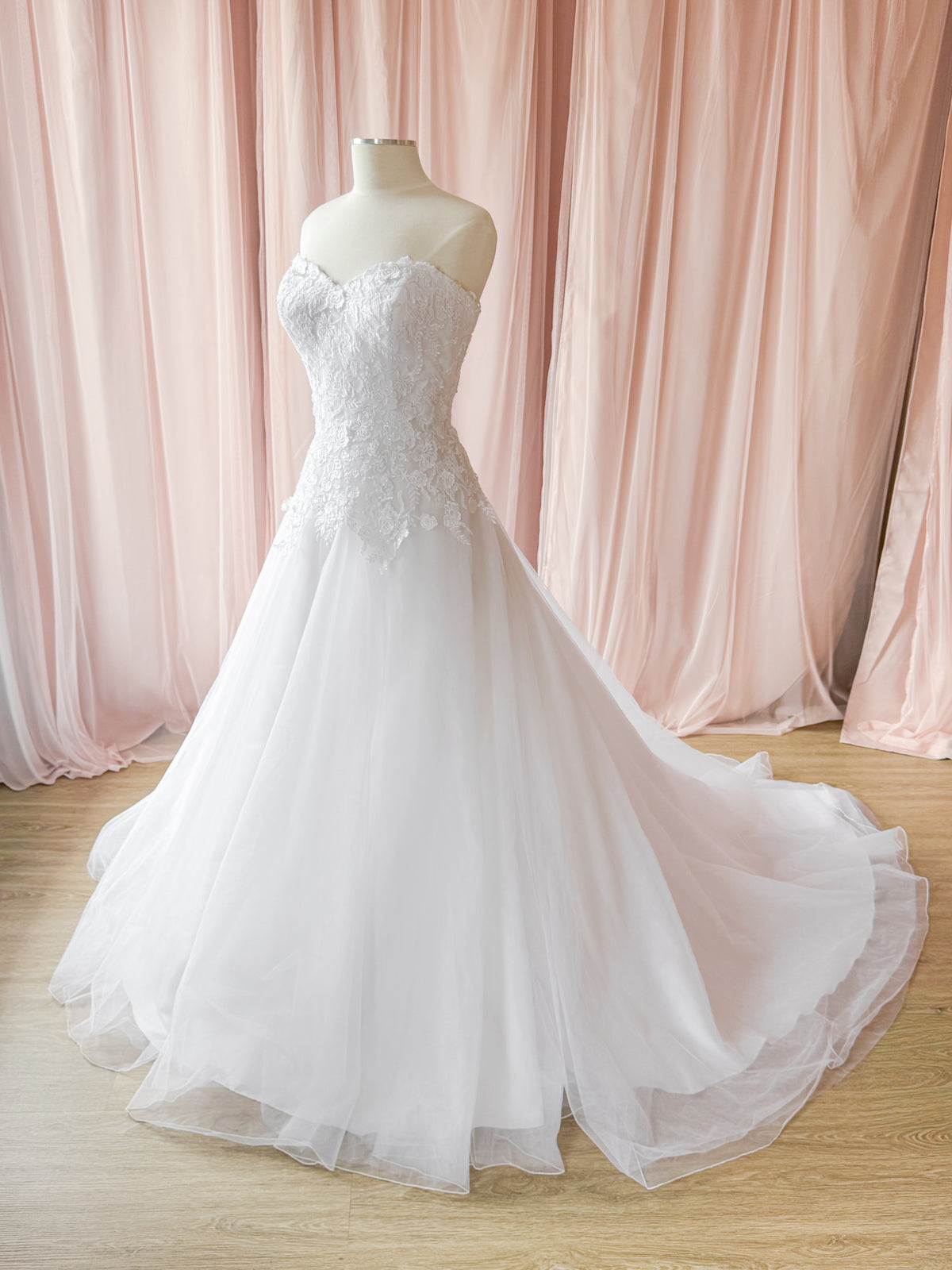 A strapless white wedding gown with a lace-embellished bodice and a full tulle skirt, displayed on a mannequin in front of soft pink draped fabric and a light wood floor.