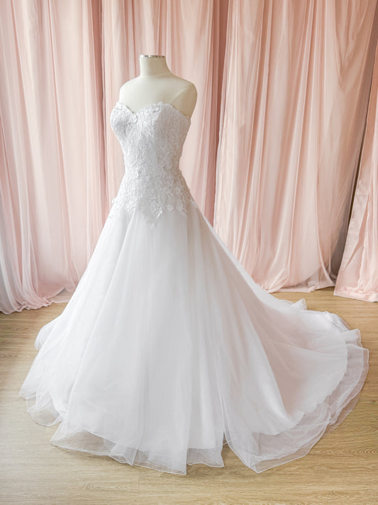 A strapless white wedding gown with a lace-embellished bodice and a full tulle skirt, displayed on a mannequin in front of soft pink draped fabric and a light wood floor.