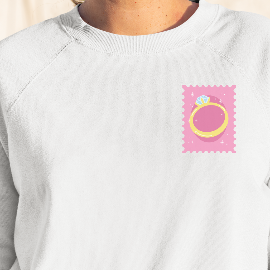 White crewneck with pink stamp and ring on top right of sweatshirt