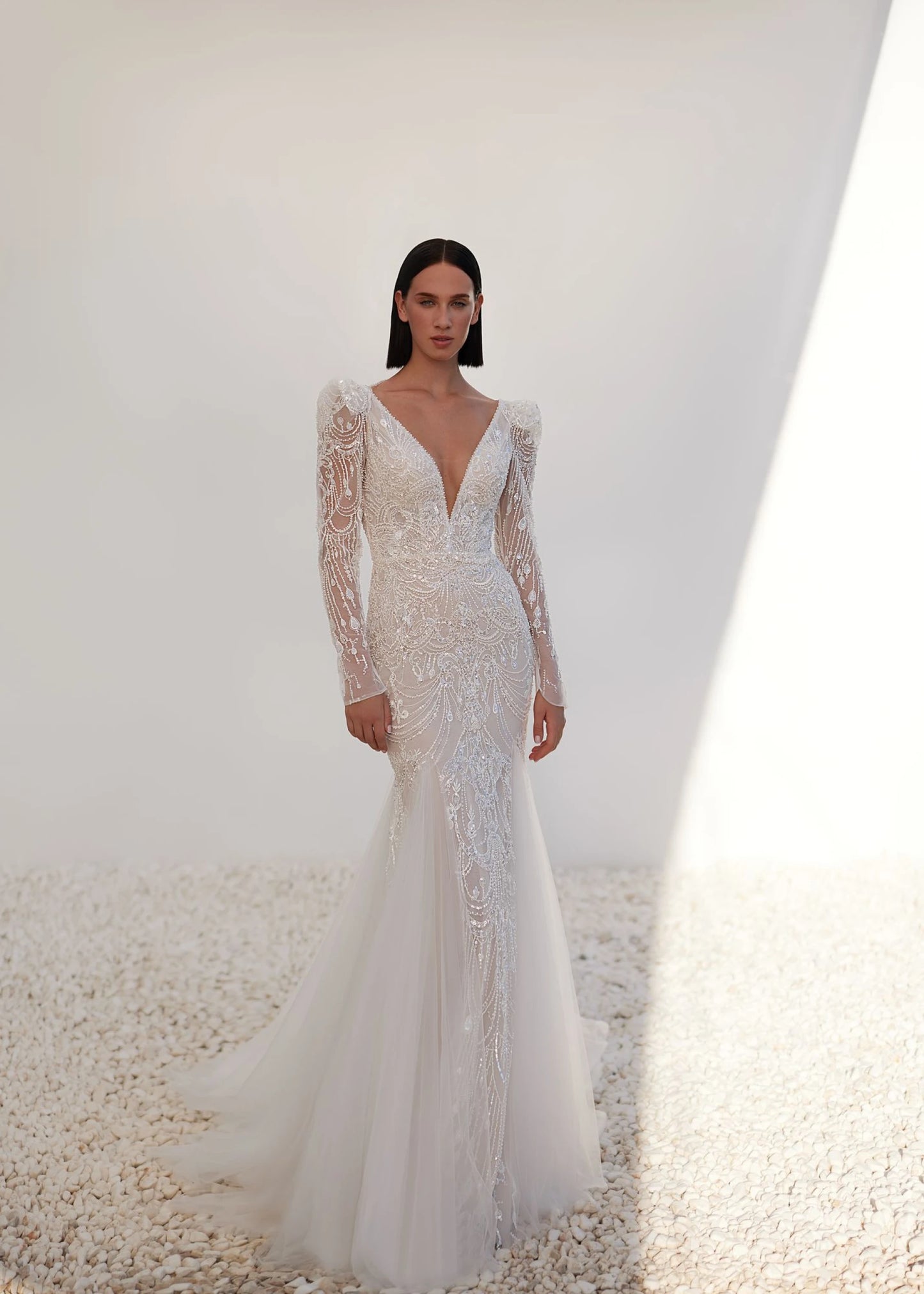 Model wearing a white wedding dress with long sleeves and a plunging neckline on a neutral background.