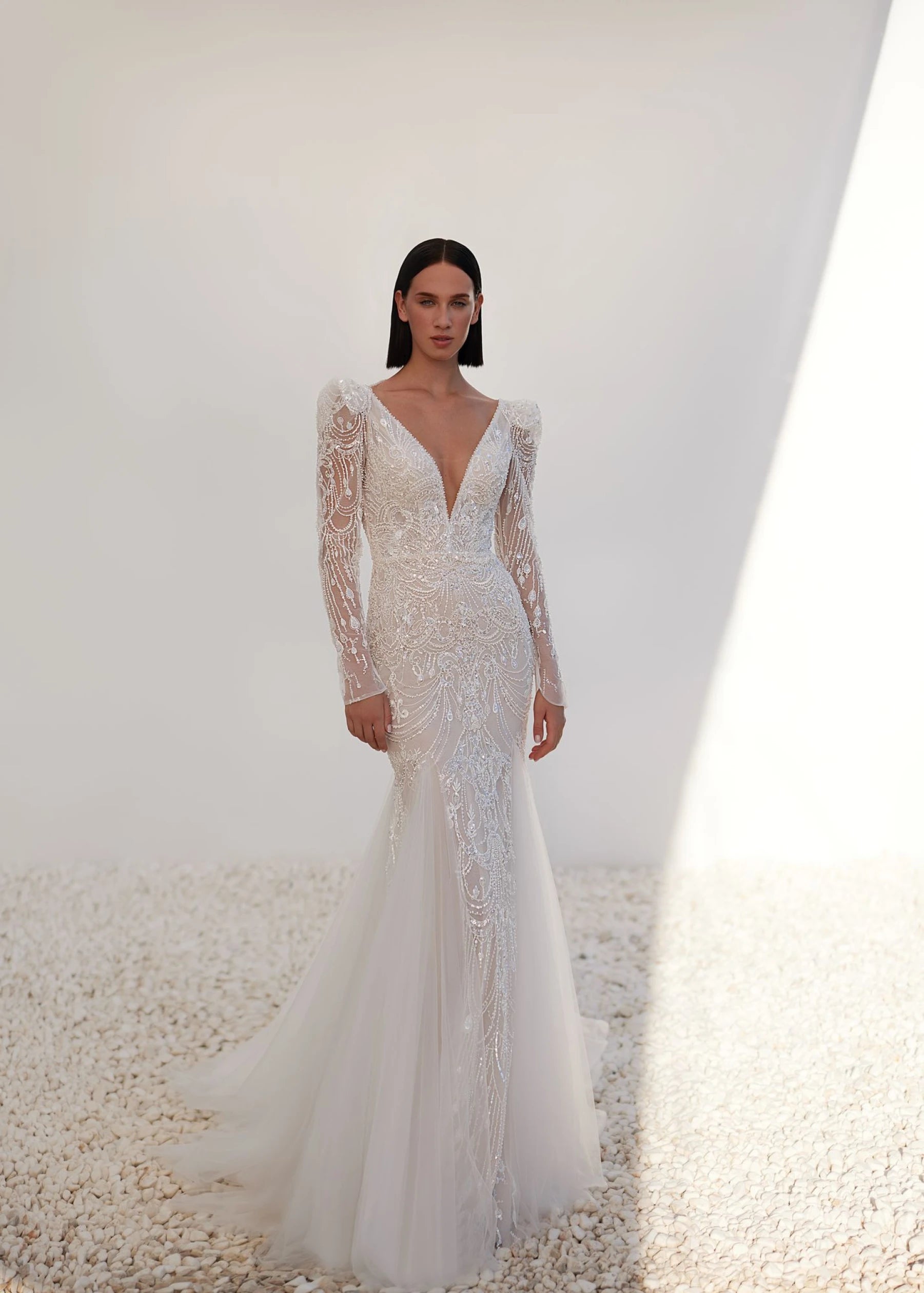 Model wearing a white wedding dress with long sleeves and a plunging neckline on a neutral background.