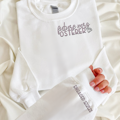 White crewneck folded with "future mrs osterer" embroidered in pink and a hand with "engaged 12.21.21" embroidered on the sleeve.