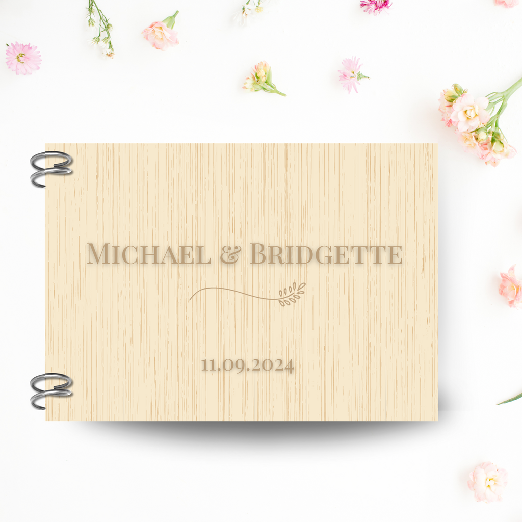 A natural wood hardcover guest book on a pink, floral background. "Michael & Bridgette, 11.09.2024" is engraved with a delicate floral design.