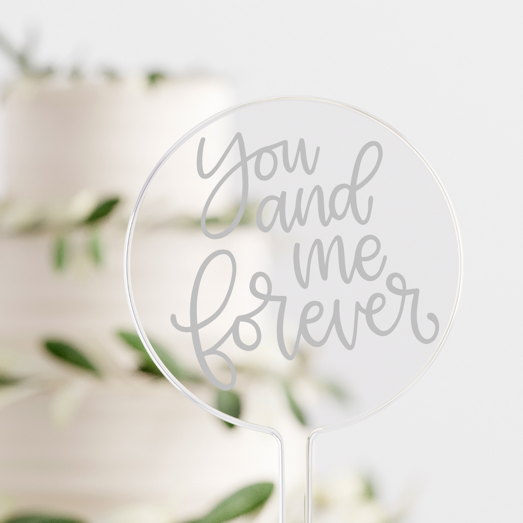 Circular acrylic cake topper with "you and me forever" written in silver