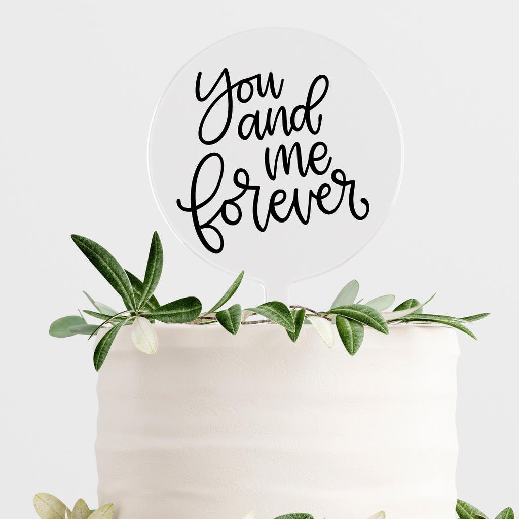 Circular acrylic cake topper with "you and me forever" written in black on simple white cake